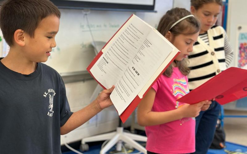 three children reading through script in front of classroom